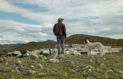 Adult man in cowboy hat and his dog standing on field with town in background against sky