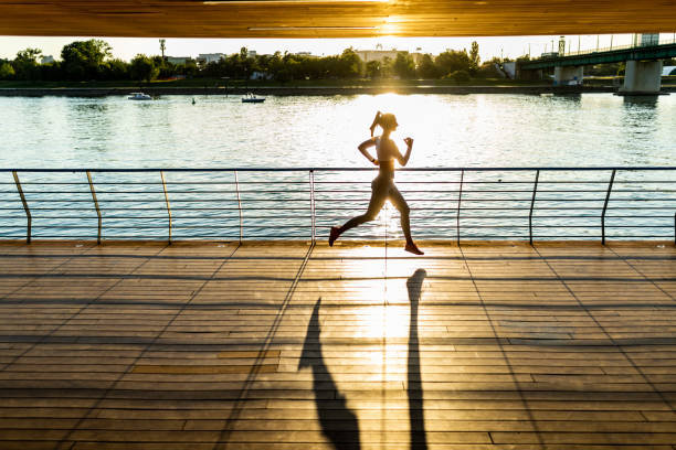 Young woman jogging and training outdoors on a wooden platform next to the river in city. Wearing sports clothing and sneakers. Setting sun in background makes beautiful light