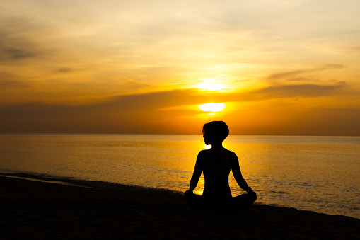 Silhouette of beautiful short hair woman sitting cross legged, meditating and practicing yoga asana in easy seat pose on the beach with yellow twilight sky sunrise in relaxation, harmony and self-love.