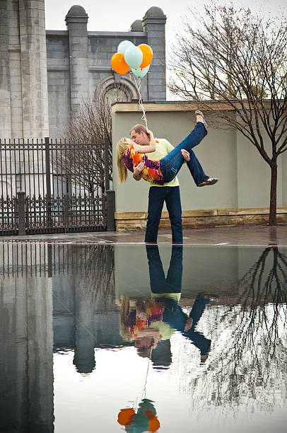 Man holding a woman and balloons while kissing her A young man is holding a young woman in front of a reflection pond.  He is also holding balloons that match her shirts with a vibrant print on it.  The are standing in front of the Salt Lake City temple in Utah.  The granite section behind them looks like a castle.  There is a perfect symmetrical reflection of the couple in the pond.  The young lady is kicking her leg out while he holds her.  It was spring and the trees don't have their leaves yet, but they have a few blossoms. mormon woman photos stock pictures, royalty-free photos & images