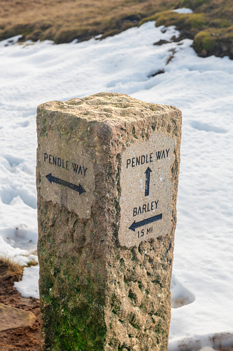 Snow and footprints around stone waymarker on the Pendle Way hiking trail at the top of Pendle Hill in Lancashire, England.