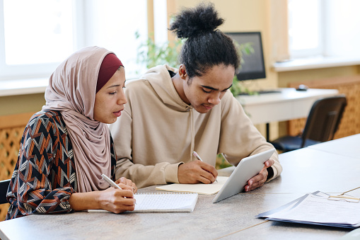 Young Black man and Middle-Eastern woman in hijab watching something on digital tablet and making notes during lesson