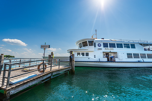 Bardolino, Italy - May 26, 2021: A white ferry boat is arriving at ferry terminal of the small Bardolino village on Lake Garda (Lago di Garda) on a sunny spring day, Verona province, Veneto, Italy, Europe. The ferry crew prepares to dock at the wooden pier on a sunny spring day.