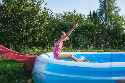 Staycation at home. Little girl in swimsuit rolls down slide into inflatable rubber pool. Swim activity in backyard of country house. Summer,relaxation on isolation,quarantine of coronavirus covid-19.