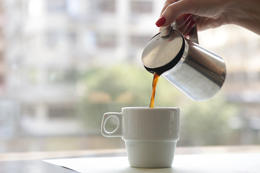 Woman's hand holding a coffee mug and pouring it into the coffee cup. Morning routine lifestyle concept, close up
