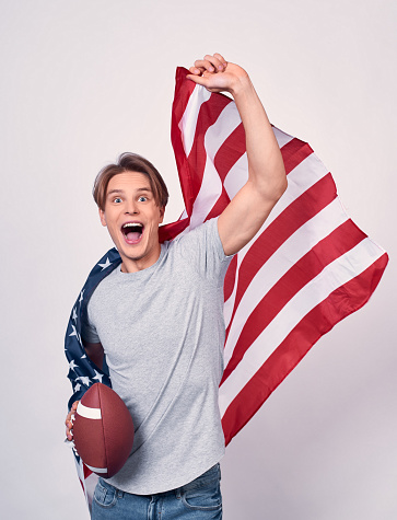Vertical shot, a young slender guy, in casual clothes, holds the American flag and ball for American football, runs and is smiling, screaming with joy. Emigrant concept, American symbolism, July 4th.