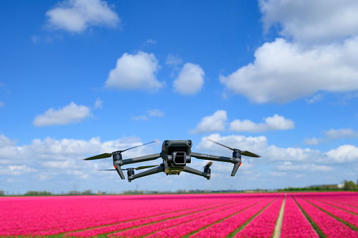 DJI Mavic 3 drone flying in the air over a field of pink tulips with the Hasselblad gimbal camera pointing forward. The Mavic 3 is a teleoperated quadcopter drone for aerial photography and videography mad by Chinese manufacturer DJI.