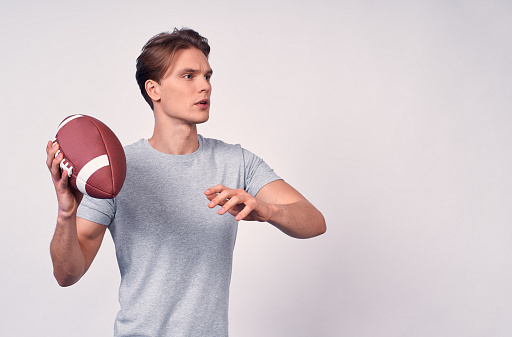 A young athletic American guy, with blond hair, in a gray basic t-shirt, holds in his right hands an American football ball, ready to make a throw. Copy space.