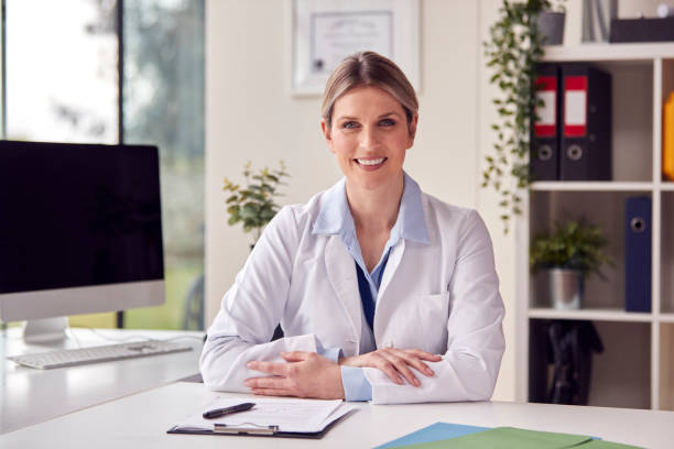 Portrait Of Smiling Female Doctor Or GP Wearing White Coat In Office Sitting At Desk stock photo