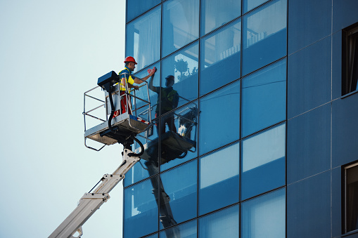Side view of a mid 30's man washing windows on a residential building facade. He's safely standing inside construction platform.