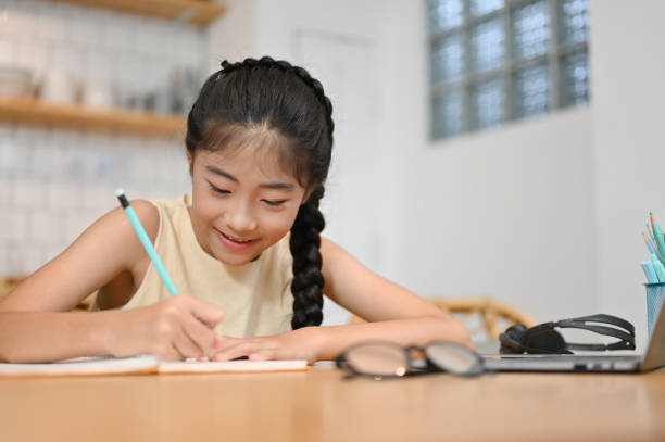 Lovely pretty Asian preteen girl doing a homework at desk Lovely pretty Asian preteen girl doing a homework at desk or writing on a school notebook in the living room. asian child studying stock pictures, royalty-free photos & images