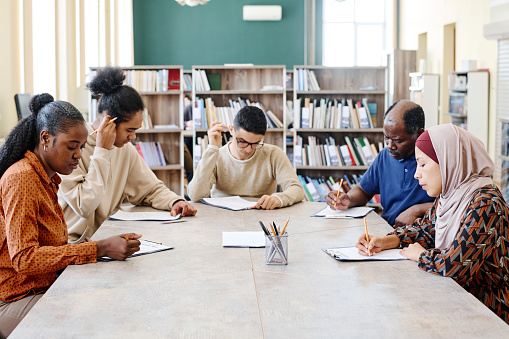 Group of ethnically diverse immigrants learning English language at school sitting in library doing grammar test