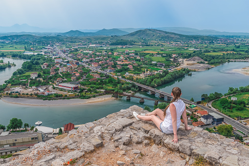 Young European woman sits on the wall of Rozafa Castle on the backdrop of Drin river valley with bridges and Bahcallek village in Albania. View from the back of a fair-haired tourist in white clothes