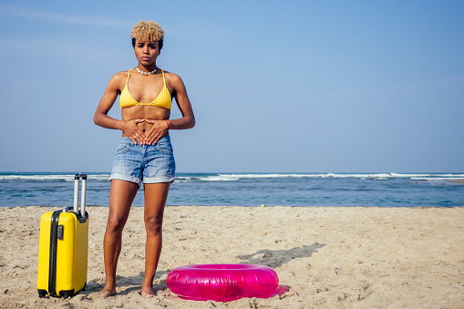 beautiful sad and sick african american woman in yellow swimsuit and denim shorts holding hands on stomach and feeling pain on beach, next to inflatable ring and suitcase on wheels...