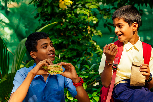 indian school boy eating sandwich and his friend with backpack sits on table and eating an apple in park.