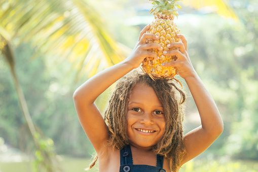 Mowgli indian boy with dreadlocks hair hiding holding a pineapple in tropics green forest background.