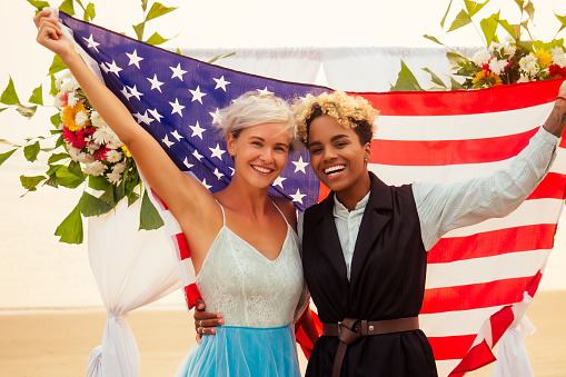 african american female groom in black suit and happy caucasian blonde bride loving together wrapped in america flag on beach ceremony under floral arch.