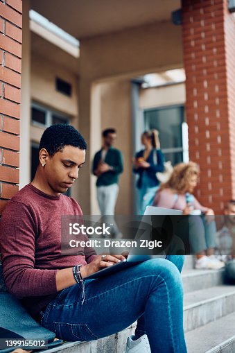 istock Young black student using laptop  while relaxing on the steps of university building. 1399155012