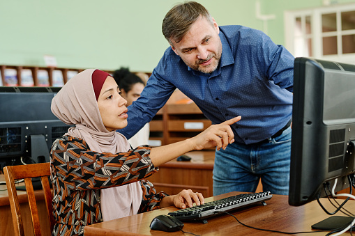 Young Muslim woman wearing hijab doing difficult task on desktop computer asking teacher to help with it