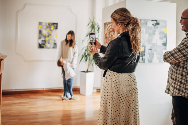 Memory from art gallery Diverse group of visitors in modern art gallery. Woman is photographing mother and daughter with smart phone. gallery opening stock pictures, royalty-free photos & images