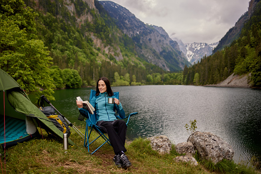 Young woman resting, sitting in camping chair and reading book by the lake