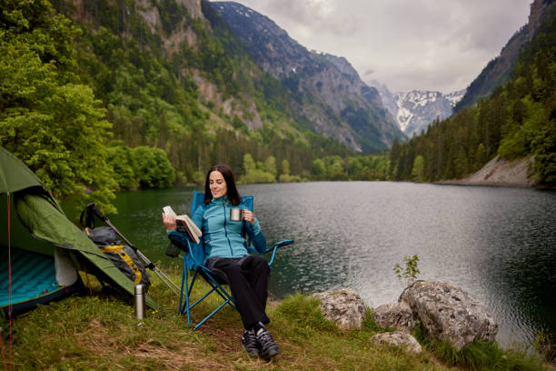 Young woman resting, sitting in camping chair and reading book by the lake stock photo