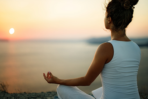 Back view of relaxed woman doing Yoga breathing exercises in Lotus position on a hill above the sea at sunrise. Copy space.