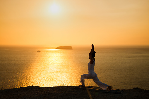 Young female athlete doing Yoga balance exercises on a hill above the sea at sunset. Copy space.