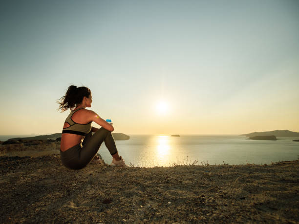 Female athlete taking a water break on a hill above the sea. Young athletic woman resting on water break on a hill above the sea at sunset. Copy space. groyne stock pictures, royalty-free photos & images