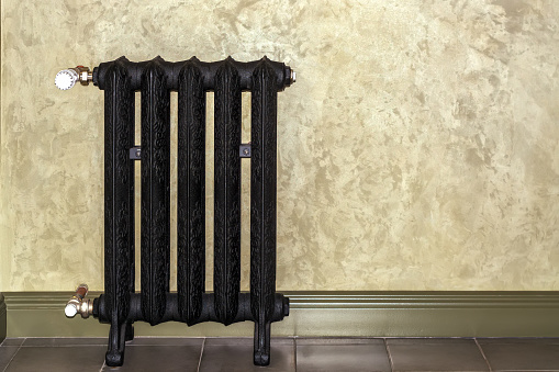 View of a black cast-iron heating radiator in the corner of a room against a beige wall.