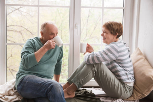 healthy seniors lifestyle. relaxing at home, Hygge. Slow life. Enjoying the little things. an elderly couple drinks tea and and communicate sitting near the window of the house stock photo