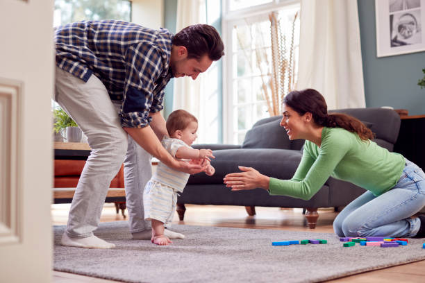 Transgender Family Encouraging Baby To Take First Steps In Lounge At Home Transgender Family Encouraging Baby To Take First Steps In Lounge At Home first steps stock pictures, royalty-free photos & images