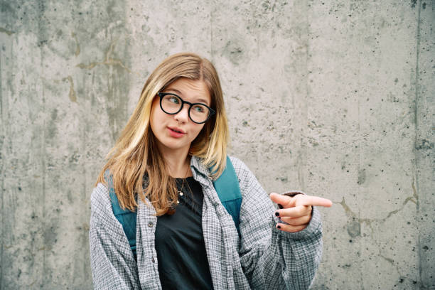 Outdoor portrait of young teenage kid girl wearing glasses and backpack, posing on grey wall background, talking and pointing finger to the side stock photo