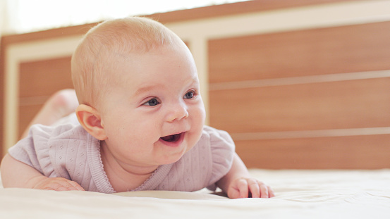 Smiling little baby girl with big blue eyes lying on her tummy on the bed at home