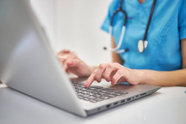 Close-up hands of unrecognizable female physician in medical uniform working typing on laptop keyboard sitting at desk stock photo