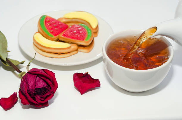 Tea is poured from a teapot into a white cup, various cookies and a dried rose on a white background. Selective focus