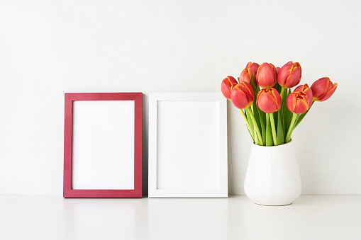 Two picture frame mockups and red tulips in a white vase on a white table by the wall. Interior, copy space for text