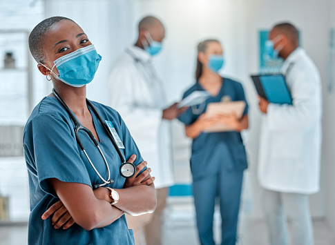 Mature african american female doctor standing with her arms crossed while working at a hospital. One expert medical professional wearing a mask while standing at work with colleagues at a clinic