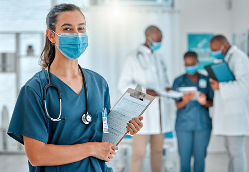 Mixed race female doctor holding a clipboard and wearing a mask while working at a hospital with colleagues. Hispanic expert medical professional ready for work at a clinic with coworkers
