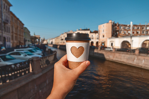 brown natural paper cup of ground coffee with carved white heart in woman's hand on background of river European city