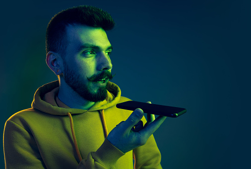 Portrait of young man in casual cloth recording voice message on phone isolated over blue background in neon light. Concept of emotions, facial expression, lifestyle, fashion, youth culture