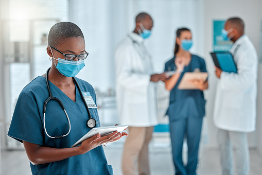 Joven doctora afroamericana trabajando en una tableta digital y usando una máscara mientras trabaja en un hospital con colegas. Enfermera negra investigando en una tableta digital en el trabajo en una clínica photo