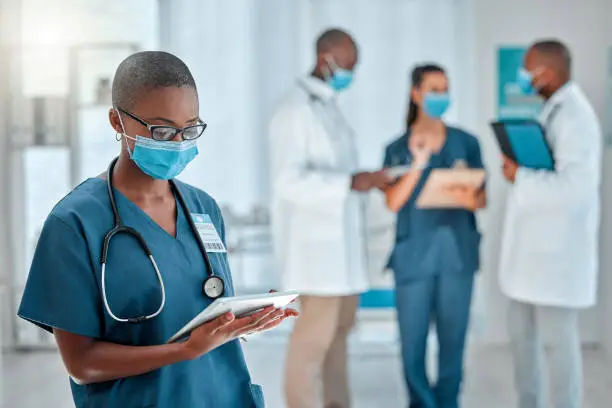 Photo of Young african american female doctor working on a digital tablet and wearing a mask while working at a hospital with colleagues. Black female nurse doing research on a digital tablet at work at a clinic