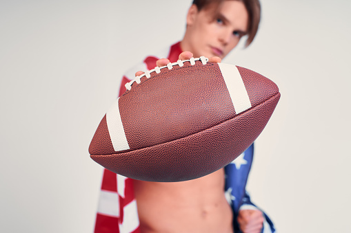 Close-up, a male athletic sexy guy with a naked torso, on his shoulders an American flag, holding a soccer ball, an American student, a football player. Focus on the ball.