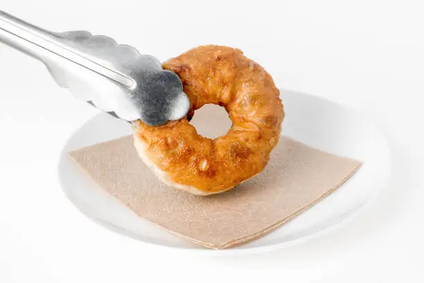 Pastry tongs hold a freshly cooked fried homemade donut and put it on a plate with a napkin. Brown fried doughnut. Yeast dough product, sweet pastries.