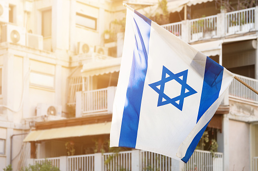 Israeli flag with star of David waving in the wind in front of windows of a residental building with sunny rays