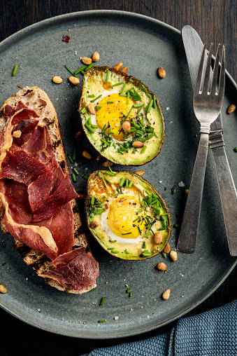 Overhead view of a dish of baked eggs with avocado’s, toasted sourdough bread and crispy serrano ham. Recipe, cut the avocado in half and remove the stone, use small sized eggs, discard some of the egg white if desired and pour the remaining yolk and white into the hole left by the avocado stone, season with salt and pepper and some chives and or some chilli flakes and in this case some roasted pine nuts. Bake for around 10-12 minutes at 200 degrees until the egg whites have set but the yolks are still runny. Serve with toasted bread, delicious.
