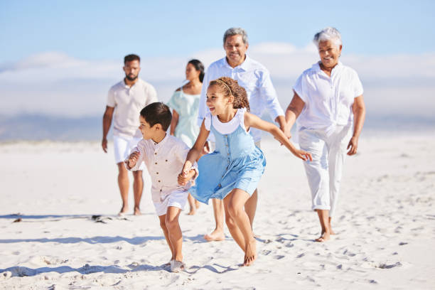istock Little girl and boy holding hands while running together on sandy beach while parents and grandparents follow in the background. Carefree sibling brother and sister having fun during summer beach vacation with their family 1399190157