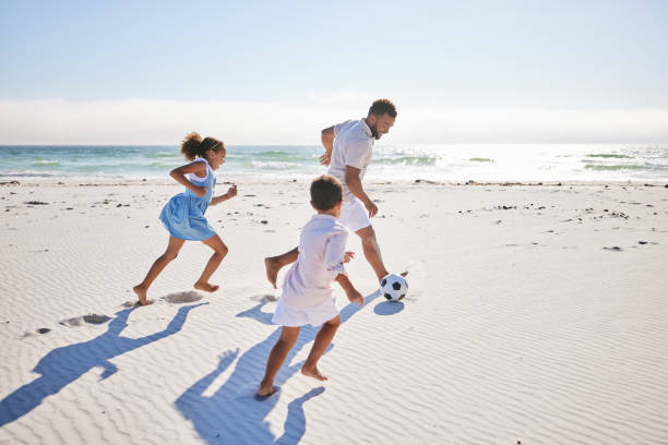 healthy father and two children playing soccer on the beach. single dad having fun and kicking ball with his daughter and son while on vacation by the sea - beach football imagens e fotografias de stock