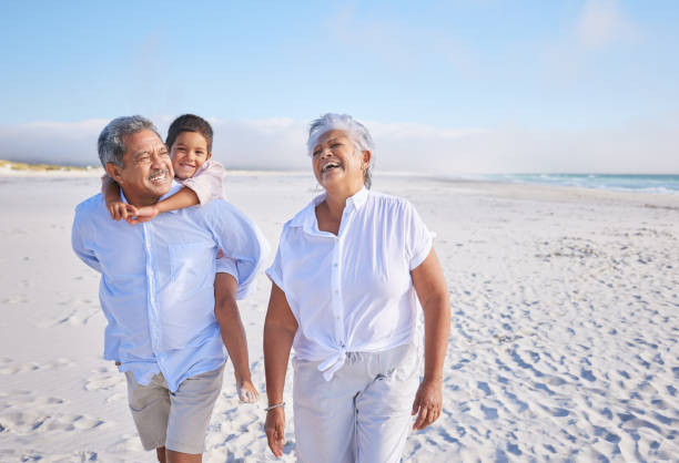 felici nonni di razza mista che camminano sulla spiaggia con il loro nipote. ragazzino che si gode un giro in piggyback sui suoi nonni durante le vacanze estive sulla spiaggia - grandfather adult affectionate little boys foto e immagini stock